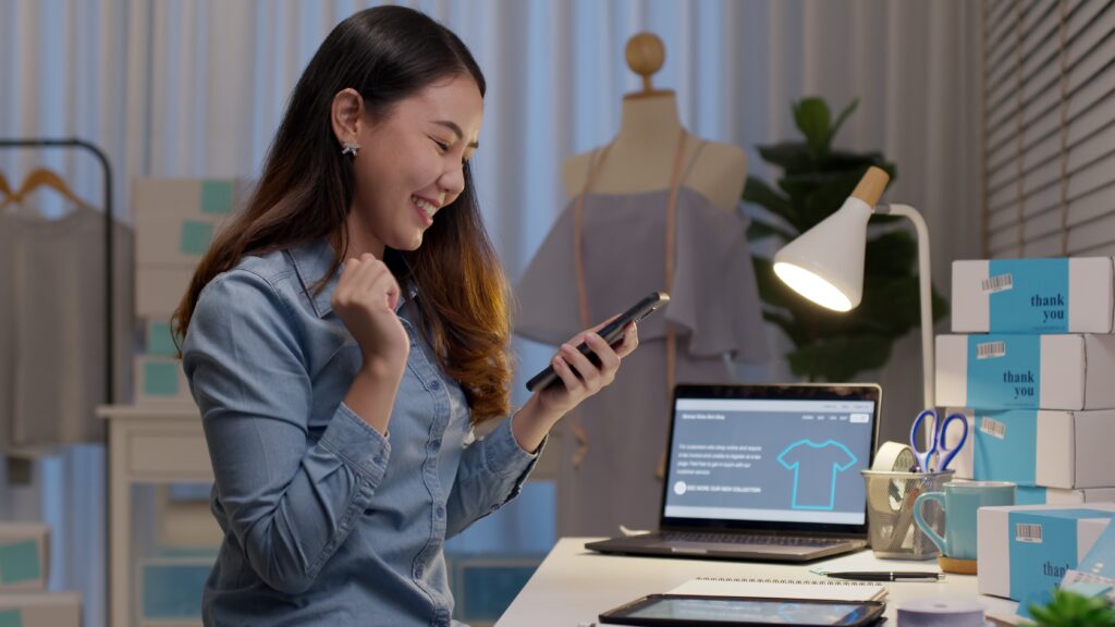 Woman celebrating success while using a smartphone in a workspace with a laptop, stationery, and neatly stacked boxes, reflecting themes of finance broking and online business management.