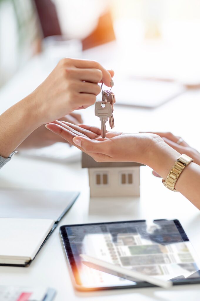 Hands exchanging house keys over a table with a miniature house model and a tablet, symbolizing mortgage transactions and home buying processes.